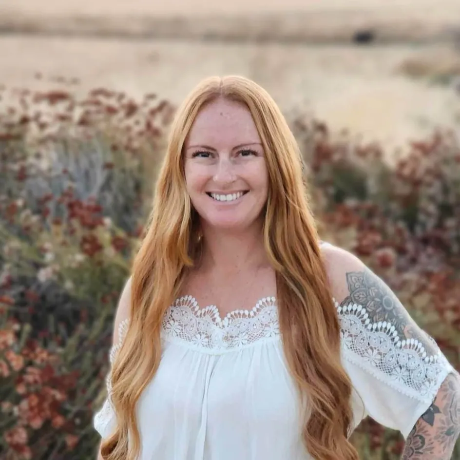 A picture of Julia Maze with long red hair, smiling outdoors in a field of dry wildflowers. She is wearing an off-the-shoulder white blouse with lace details and has visible tattoos on her arm. The background is softly blurred, emphasizing the natural landscape behind her